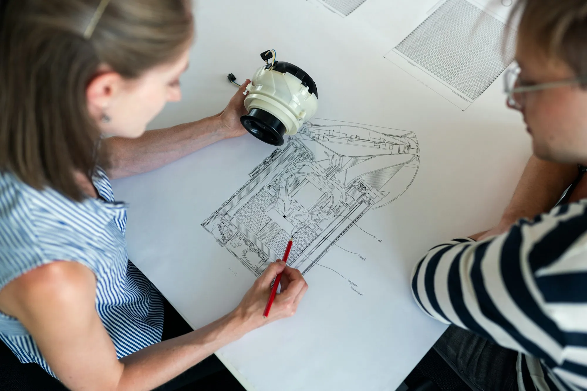Two people reviewing architectural blueprints together on a large format drawing, one pointing with a red pencil at structural details